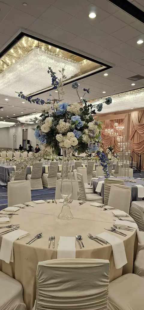 Blue and ivory floral centerpiece on a tall crystal stand above a round table, with a ballroom of ivory chairs behind.