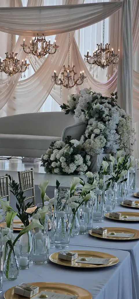 Long head table with gold chargers, calla lilies in glass bud vases and gold chiavari chairs, before a white lounge sofa, white hydrangeas and blush drapery hung with crystal chandeliers.