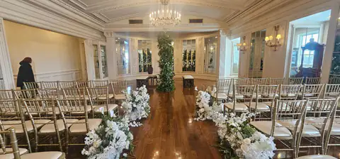 Ceremony aisle in a white paneled hall, lined with gold chiavari chairs and low clusters of white orchids and roses, leading to a column of greenery beneath a crystal chandelier.