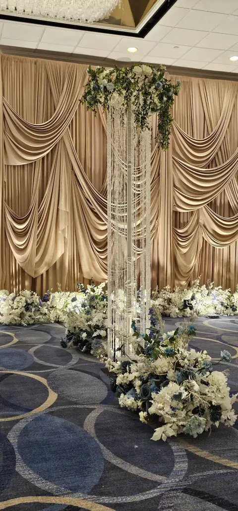 Champagne swag drapery behind a crystal-strand pillar crowned with white and blue florals, with flowers massed along the floor.