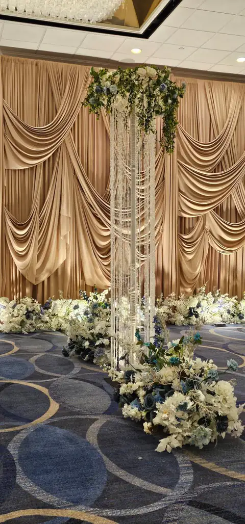 Champagne swag drapery behind a crystal-strand pillar crowned with white and blue florals, with a low line of flowers along the stage edge.
