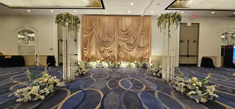 Wide view of a sculpted champagne fan-pleat stage backdrop edged with white and blue florals, flanked by crystal-strand pillars topped with floral arrangements.