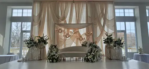 Reception stage in a bright hall with tall windows: ivory swag drapery, crystal chandeliers, a white curved sofa, white rose arrangements and gold candelabras.