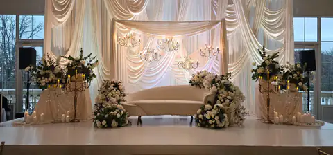 Ivory stage with a sculpted sunburst pleat backdrop, crystal chandeliers, a draped canopy and gold candelabras wrapped in white florals.
